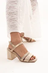 Sandalia de cuero con acabado metalizado dorado, diseño de doble tira delantera y cierre con hebilla ajustable en el tobillo. Cuenta con taco cuadrado y puntera cuadrada.