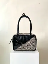 Cartera rígida negra con detalles en rojo y tachas plateadas. Tiene herrajes y cierre metálico plateado marca YKK, asa fina para llevar al hombro o en la mano y correa larga extra regulable. El interior es forrado, tiene un bolsillo con cierre enmarcado en cuero, y un segundo bolsillo plaqué.