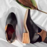 Botín de cuero vacuno color negro, con diseño de corte lateral, cierre metálico en el talón y taco foliado de madera en tono natural.