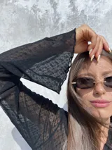 Remera de encaje negro con diseño cruzado frontal y lazo lateral para ajustar. Presenta mangas largas con terminación acampanada y un delicado patrón de pequeñas flores bordadas en toda la prenda.