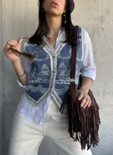 Chaleco de jean azul con bordados florales blancos y ribete de encaje. Cierre frontal con botones.