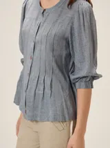 Camisa de lino mezcla color beige, con cuello redondo, cierre frontal con botones y mangas tres cuartos con puños elásticos. Presenta detalles de pespuntes verticales en zigzag en el frente.