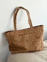 Cartera tipo tote color tostado, hecha en cuero graneado con textura uniforme y suave al tacto. Tiene manijas de 55 cm de largo, un bolsillo frontal con broche imantado y un bolsillo trasero con cierre metálico. El compartimento principal cierra con cierre metálico y el interior está forrado con tres bolsillos: uno con cierre, uno para celular y otro tipo sobre con solapa y botón.