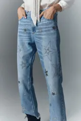 Jean de tiro alto en denim azul rígido, con lavado localizado, bigotes y apliques de estrellas brillantes. Presenta pierna recta y ruedo desflecado.