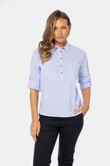 Camisa blanca de manga larga con cuello y tapeta de medio botón. Presenta dos bolsillos frontales con tapa y botones metálicos. Las mangas tienen presillas para sujetar el puño enrollado.