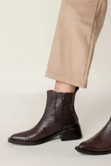 Bota tipo chelsea de caña media con acabado efecto cocodrilo en color chocolate. Presenta paneles elásticos laterales, puntera cuadrada y taco ancho geométrico.