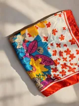 Pañuelo cuadrado de seda con estampado floral multicolor que combina tonos rojos, amarillos, azules y fucsias sobre fondo blanco, con borde rojo.
