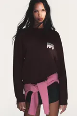 Camiseta de manga larga color marrón oscuro con efecto lavado. Presenta un cuello redondo y un estampado en el pecho con el texto "NEW YORK" en verde y "POP UP" en rosado.