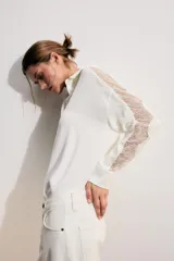 Camisa blanca de manga larga con cuello solapa, cierre frontal con botones y bajo asimétrico. Presenta detalles de puntilla combinada a tono en las mangas y hombros.