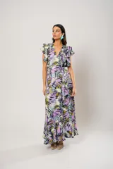 Vestido largo con estampado floral en tonos lila, verde y negro. Tiene escote en V, mangas cortas con volados y corte cruzado con lazo en la cintura.