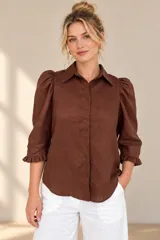 Camisa de lino color blanca, con cuello clásico, mangas tres cuartos abullonadas en los hombros y terminación con volados en los puños, cierre frontal con botones ocultos.