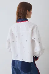 Camisa de lino blanca con estampado de motivos deportivos, cuello y puños tejidos a mano en crochet azul con borde rojo. Presenta cierre frontal con botones y un bolsillo en el pecho.