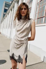 Vestido corto de cuello subido y sin mangas, con diseño fluido y pliegues en los hombros. Presenta un detalle de frunces en el frente, una abertura lateral y un borde inferior decorado con encaje negro en contraste.