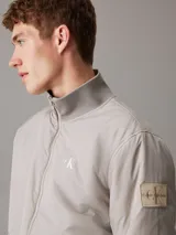 Campera Calvin Klein color gris claro, confeccionada en nailon 100% reciclado. Tiene cuello alto de canalé, puños y bajo elásticos, cierre de cremallera, bolsillos en la cintura, forro ligeramente acolchado y logo de la marca en el pecho y la manga.