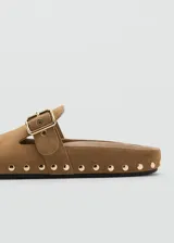 Mocasines destalonados de piel bovina color marrón con tachas metálicas doradas alrededor de la suela y una tira en el empeine con hebilla ajustable.