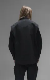 Chaqueta tipo overshirt de microfibra, color negro, con calce oversize y ruedo curvo. Presenta cierre frontal con broches a presión, bolsillos frontales de gran tamaño y cierres laterales a lo largo de las mangas para ajustar su amplitud.