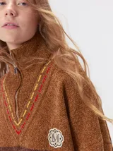 Pullover de punto con textura aterciopelada, diseño oversize, cuello polo con cierre frontal parcial y detalles de pespuntes en contraste. Presenta rayas horizontales en tonos tierra y un parche bordado con la letra M en el pecho.