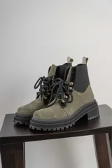 Borcego de gamuza color chocolate con paneles laterales elásticos negros y suela track dentada. Presenta sistema de cordones con ganchos metálicos y tirador en la parte trasera.