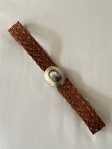 Cinto trenzado de cuero color marrón, con hebilla metálica ovalada grabada y detalle central de piedra natural.