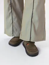 Slipper tipo guillermina confeccionada en gamuza color marrón, con plataforma de goma negra y doble hebilla ajustable. Presenta un pespunte decorativo en el borde inferior y cuenta con interior recubierto en cuero.