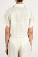 Camisa crop fluida de manga corta color crema con cuello camisero, botonadura frontal oculta y detalle de nudo en el frente.
