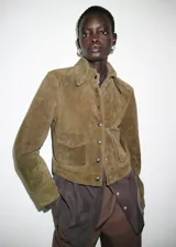 Chaqueta corta de piel de serraje color marrón, con diseño recto, cuello camisero, manga larga y cierre frontal de botones metálicos. Cuenta con dos bolsillos de parche con solapa en la parte delantera y forro interior.