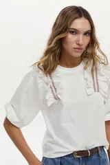 Blusa blanca con cuerpo de jersey y mangas de tela plana abullonadas. Cuenta con volados en los hombros decorados con bordados en contraste.