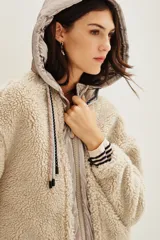 Campera de estilo híbrido con cuerpo de corderito sintético color beige y panel frontal central con cierre y capucha de tela técnica metalizada en tono grisáceo. Presenta cordones ajustables en la capucha con diseño estampado y puños con detalle de rayas.