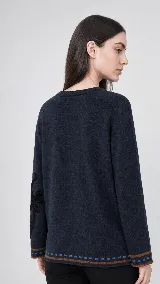 Sweater de lana color beige con calce holgado y cuello redondo. Presenta un bordado negro con diseño de arabescos en una de las mangas y detalles de rayas y puntos en contraste en el ruedo y los puños.