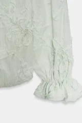 Blusa de escote pico y manga larga con puño elástico y volante. Presenta un diseño de rayas verticales finas en azul claro y blanco, con flores en relieve.