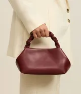 Cartera pequeña color bordó, con asa de mano trenzada y estructura rígida de líneas redondeadas.