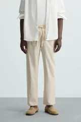 Pantalón de corte relajado color beige confeccionado en tejido de mezcla de algodón y lino. Posee cintura elástica ajustable con cordón, bolsillos frontales y bolsillos traseros de vivo.