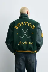 Cazadora estilo varsity con cuerpo de mezcla de lana en color verde oscuro y mangas de efecto piel a tono. Presenta cuello solapa, cierre frontal con botones a presión, bolsillos laterales de vivo y puños y bajo acanalados con rayas amarillas. Incluye bordados y parches decorativos en el frente con la inscripción Boston Archive.