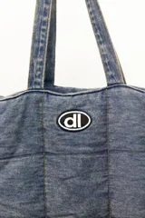 Bolso tipo shopper confeccionado en denim acolchado con diseño de paneles cuadrados. Presenta doble asa de hombro y un parche ovalado con el logo 'dl' en la parte frontal.