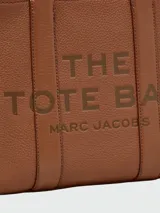 Bolso tipo tote de cuero negro, modelo The Leather Medium Tote Bag de Marc Jacobs. Presenta doble asa superior, correa ajustable para el hombro y el texto grabado "THE TOTE BAG MARC JACOBS" en el frente.