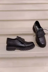 Mocasín de cuero vacuno negro con textura tipo reptil, suela track de goma, cordones finos y solapa con flecos sobre el empeine.