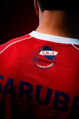 Camiseta de fútbol Umbro del Club Nacional de Football, modelo Away 1. Diseño principal en color rojo con cuello en V azul, detalles blancos en los hombros y puños blancos. Incluye el escudo del club, logo de Umbro y sponsors estampados.