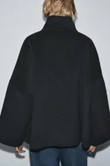 Cazadora de corte amplio confeccionada en hilatura con lana, color beige. Presenta cuello subido, manga larga y cierre frontal con botones negros. Destaca por sus acabados con costuras de hilo a contraste en los bordes.