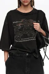 Remera corta de corte oversize color negro, confeccionada en mezcla de algodón. Presenta cuello redondo, mangas cortas amplias y un gran bordado frontal con diseño estilo etiqueta y texto.