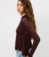 Blusa de encaje color bordó con trama floral y transparencia. Presenta cuello medio, mangas largas ajustadas y un recorte debajo del busto que genera un efecto peplum con volados en el dobladillo.