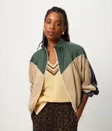 Campera rompeviento de diseño tricolor con bloques de color en verde, beige y blanco. Presenta cuello alto, cierre frontal con cremallera, mangas raglán y puños ajustados. El ruedo incluye un cordón regulable para un ajuste personalizado.