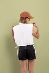 Camisa blanca sin mangas de lino y algodón, con cuello clásico, cierre frontal con botones, dos bolsillos con solapa en el pecho, hombreras y cordón ajustable en la cintura.