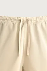 Bermuda de corte relajado confeccionada en tejido de algodón, color beige con vivos en contraste color verde oscuro. Cuenta con cintura elástica ajustable con cordón, bolsillos frontales y bolsillos traseros de plastrón.
