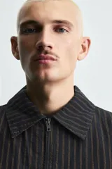 Cazadora de hombre de ajuste cropped fit, confeccionada en tejido de algodón con patrón de rayas verticales finas en tonos oscuros (negro/azul marino y marrón/beige). Presenta cuello solapa, cierre frontal de cremallera, bolsillos laterales y elástico en puños y bajo.