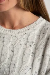 Sweater de punto color blanco roto con efecto tweed (pequeñas motas negras), cuello redondo y manga larga acampanada. Presenta un tejido texturado con trenzas verticales.