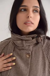 Chaqueta de corte estructurado con estampado cuadrille en tonos marrones y beige. Presenta cuello alto con tira ajustable, cierre frontal con botones, charreteras en los hombros y puños con detalle de hebilla.