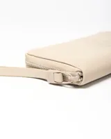Billetera rectangular Extra Large de símil cuero color taupe, con cierre perimetral y tira corta para la muñeca. Presenta un aplique circular con el logo de la marca grabado en el centro.