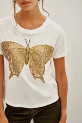 Remera de algodón color blanco con cuello redondo y mangas cortas con dobladillo. Presenta un bordado detallado de una mariposa en tonos dorados en la parte frontal.