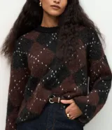 Suéter de punto pesado con diseño de cuadros argyle en tonos marrón y negro, con detalles de puntos claros. Presenta un corte oversize, cuello redondo acanalado y mangas largas.