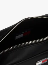 Cartera tipo bandolera de diseño compacto y minimalista, confeccionada en poliuretano con acabado liso. Presenta cierre de cremallera superior y una insignia metálica distintiva de Tommy Jeans en el centro del frente.